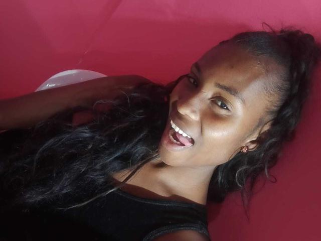 HornyCocoa with dark skin, long black wavy hair, and a happy open-mouth smile, wearing a black shirt, against a red background. Her shiny face is visible.