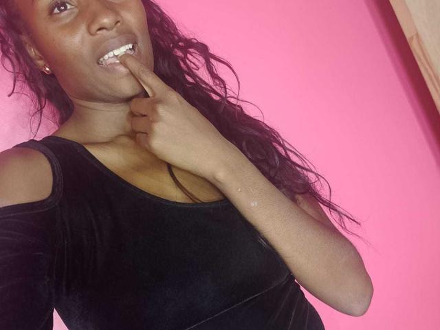 HornyCocoa with dark skin, long wavy black hair, wearing a black velvet top, biting her finger, showing white teeth, pink background, wearing small gold earrings.
