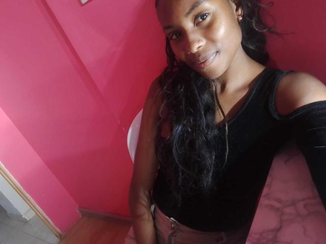 HornyCocoa with dark skin, long black wavy hair, black off-shoulder top, smiling, pink wall, wooden floor, and pink marble-like surface in the background.