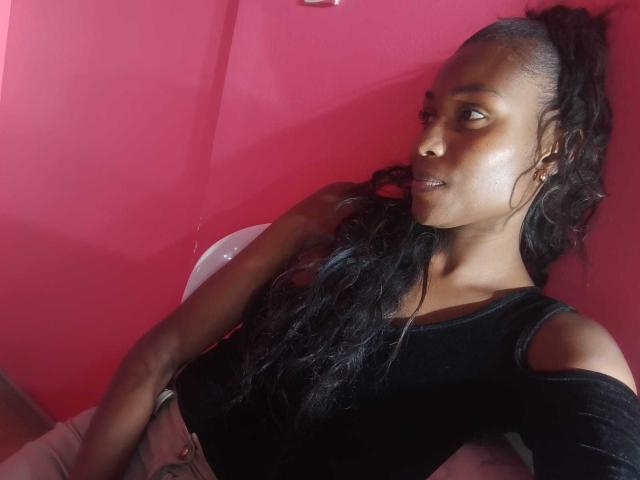 HornyCocoa with dark skin, long black hair in a ponytail, wearing a black off-shoulder top, sitting against a red wall, looking to the side, with a slight smile.