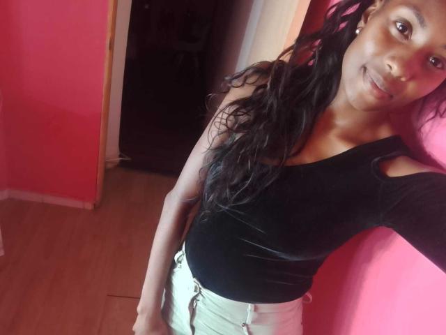 HornyCocoa has dark skin, long black wavy hair, black sleeveless top, showing small tits, and wears white high-waisted pants, standing against a pink wall, smiling.