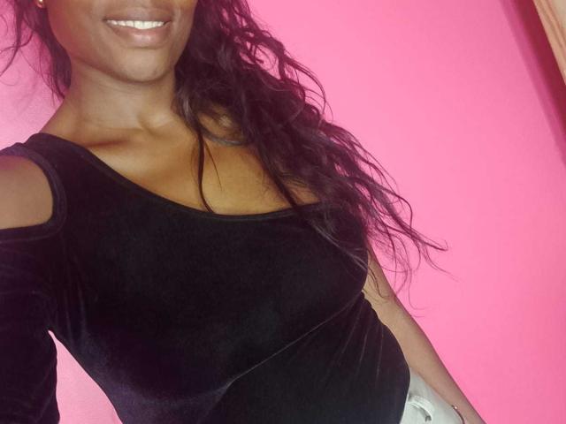 HornyCocoa, dark-skinned with wavy black hair, smiling, wearing a black velvet top with cut-out shoulders, showing her small, perky tits. Pink background.