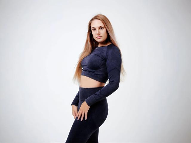 NabaPretty has long blonde hair, wearing a dark blue crop top and matching leggings, standing with hands near her ass, looking at the camera with a serious expression.