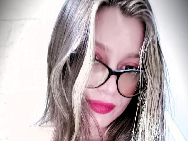 KoraSweet has blonde hair, black glasses, and red lipstick, winking with a slight smile. Her ass is not visible. Her tits are not visible. She looks cute.