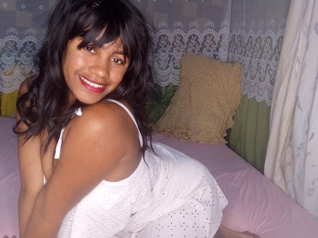 LayahCoquine a la peau brun foncé, les cheveux noirs et ondulés, et elle sourit avec du rouge à lèvres rose. Elle porte un haut blanc, orné de dentelle, sans manches, qui révèle ses petites seins fermes..