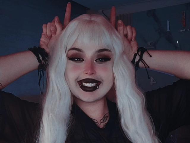 MadameRaven with white hair, dark makeup, black lipstick, and black shirt, raising her hands with fingers pointing up, smiling with a visible tongue piercing. She has tattoos.