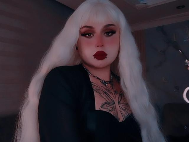 MadameRaven has long white hair, red lipstick, and a black top with a butterfly tattoo on her chest. She's wearing a chain necklace and looking at the camera.