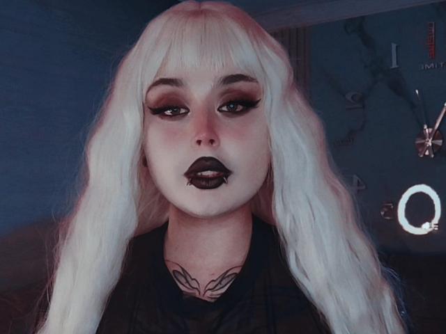 MadameRaven with platinum blonde hair, dark lipstick, heavy eyeliner, and tattoo on her neck, staring directly at the camera with a serious expression, wearing a dark shirt.