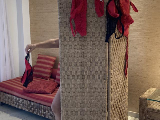 DeesseBleue's hand holds red and black lace lingerie, standing beside a woven wall with more lingerie hanging. She's partially visible, with her bare ass and red-striped bed in background.