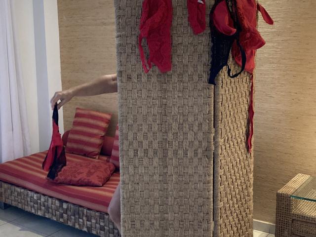 DeesseBleue's hand holding red lingerie, arm partially visible, wicker divider with more red lingerie hanging, red-striped bed with pillows in background, wicker furniture.
