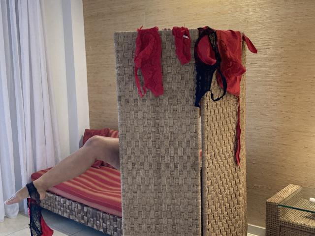 DeesseBleue's legs are on a red-striped bed, with red and black lace lingerie hanging on a woven room divider. Her bare ass is partially visible.