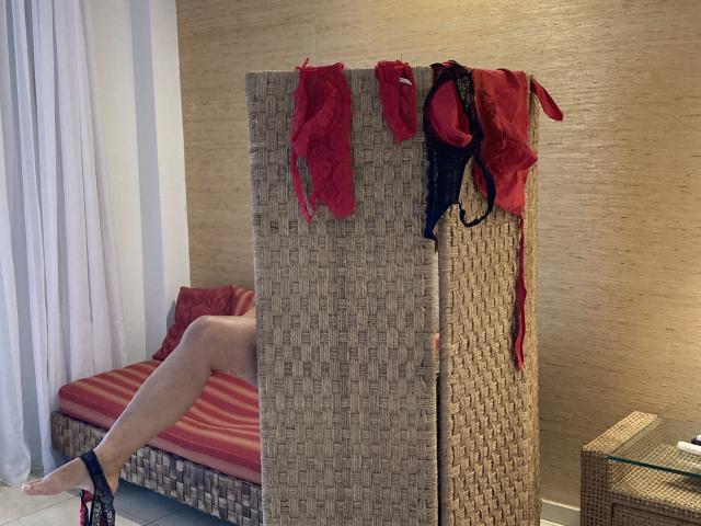 DeesseBleue's legs on a wicker bed with red pillows, wearing black heels. Red and black lingerie hangs on a woven divider. DeesseBleue's bare legs are visible.