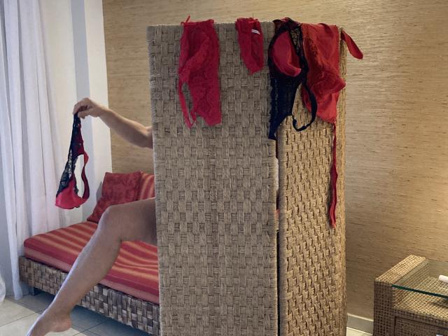 DeesseBleue's hand holds red panties, her bare leg on a wicker bed. Red and black lingerie hangs on the wicker divider. Her boobs are partially visible.