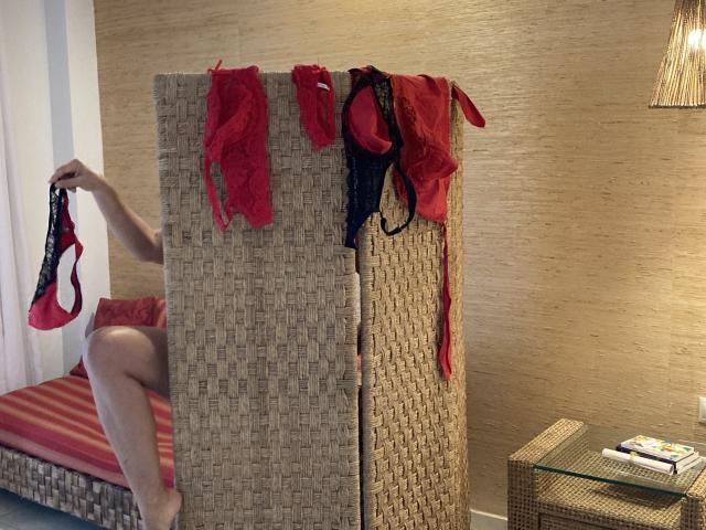 DeesseBleue's red and black lace panties and bra are draped over a woven room divider, while she holds another red bra, her bare legs and red-striped bed visible.
