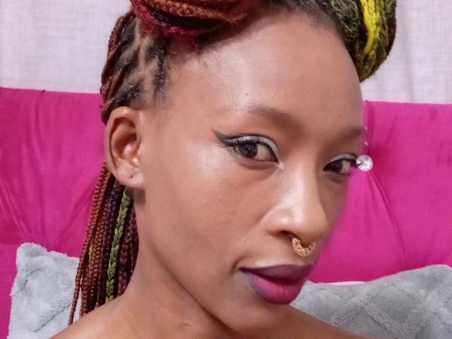 SexyBadX has brown skin, red and yellow braided hair, eyeliner, purple lipstick, a nose ring, and a small facial piercing. She's looking slightly to the side.