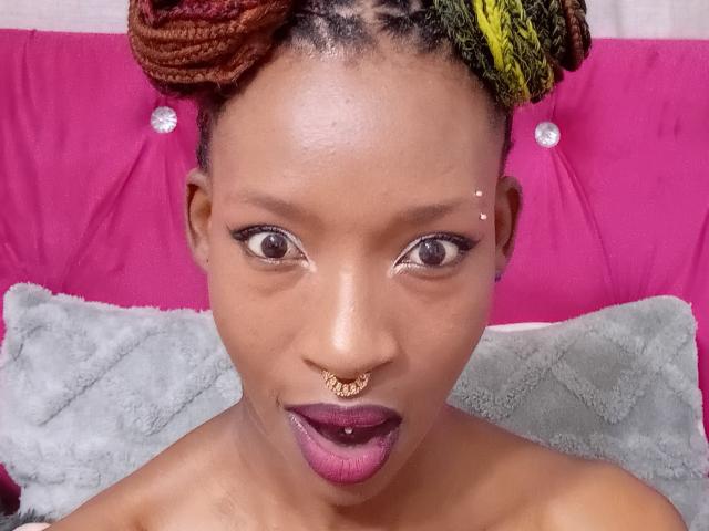SexyBadX with brown eyes, nose ring, and lip piercing, looking at the camera, wearing braided hair with red and yellow, purple lipstick, and no clothes, against a pink and gray background.