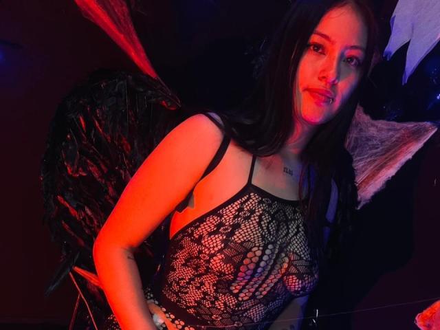 AsiaticVixen with dark hair, wearing a black mesh top, showing small tits, and black feathered wings, lit with red and blue light, smiling slightly, with a nose ring.