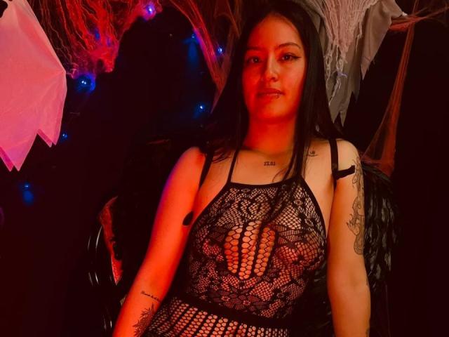 AsiaticVixen with long black hair, wearing a black lace bodysuit, exposing her tits through the mesh, has tattoos on her arm and chest, sitting in a dark, red-lit room.