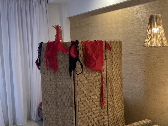 Weavy wicker room divider with red and black lingerie, including bras and panties, hanging over the top. Warm hanging light. DeesseBleue's hand is raised above the lingerie.