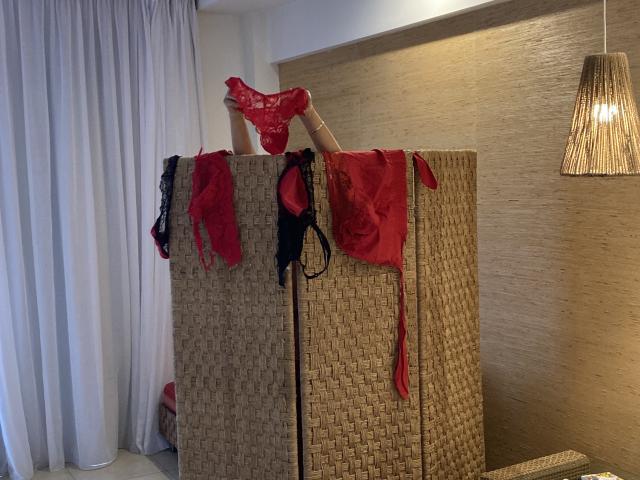 DeesseBleue's legs are up, showing off red lace panties, and black red lace bras and panties are hanging from a woven screen. Warm light, beige wall.