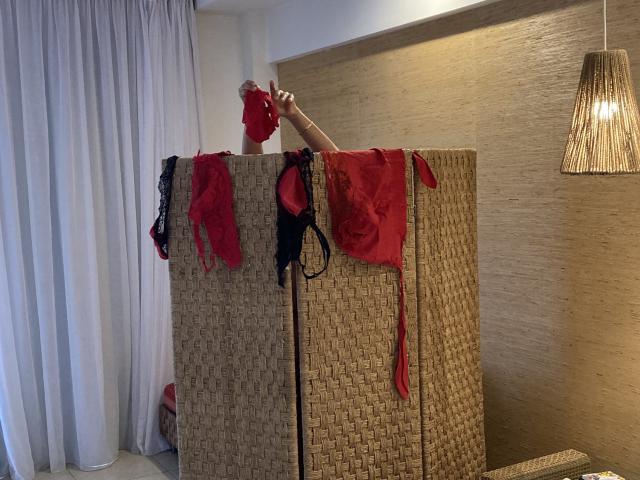 DeesseBleue stands behind a wicker screen, wearing a red mask, with red and black lingerie hanging over the top. Her arm is raised, hand near mask.