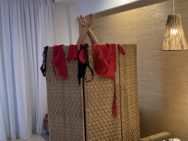 DeesseBleue's legs are raised, arms crossed, with red and black lingerie draped over a woven room divider, under a hanging light, in a softly lit bedroom.
