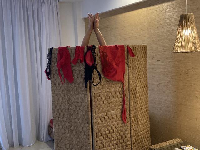 DeesseBleue's arms above a woven divider, red and black lingerie hanging, hands touching top, dim room, wicker chair, lamp, white curtains.