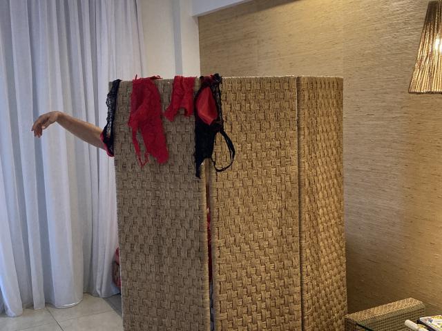 DeesseBleue's arm extends from behind a woven partition, with red and black lingerie hanging above it, including a bra and a thong, in a well-lit room.