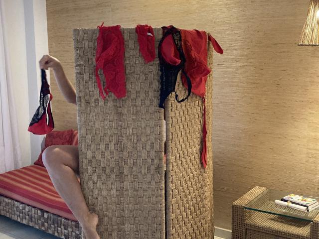 DeesseBleue's bare legs and arms hold red and black lace panties, bras, and a garter, draped over a woven room divider, next to a wicker bed and glass table.