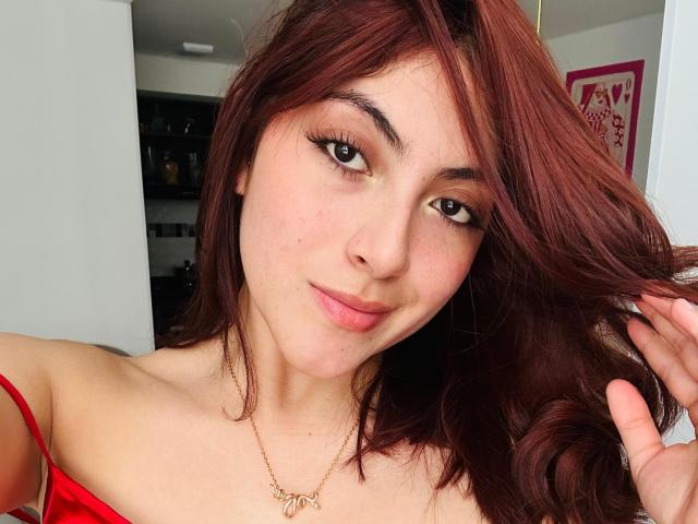SpencerLe tem cabelo ruivo, um tom de pele claro e está vestindo uma blusa vermelha com alças e um colar dourado. Seus cílios são longos e ela está segurando o cabelo..