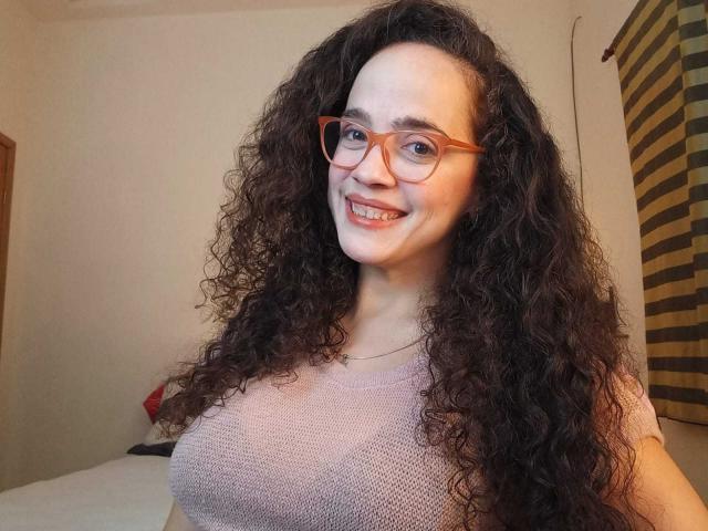 AmyFlores, met lang, krullend bruin haar, draagt een rode bril en een roze gebreide top, en glimlacht naar de camera terwijl ze haar borsten laat zien. Ze heeft een lichte huid en een schattig gezicht..