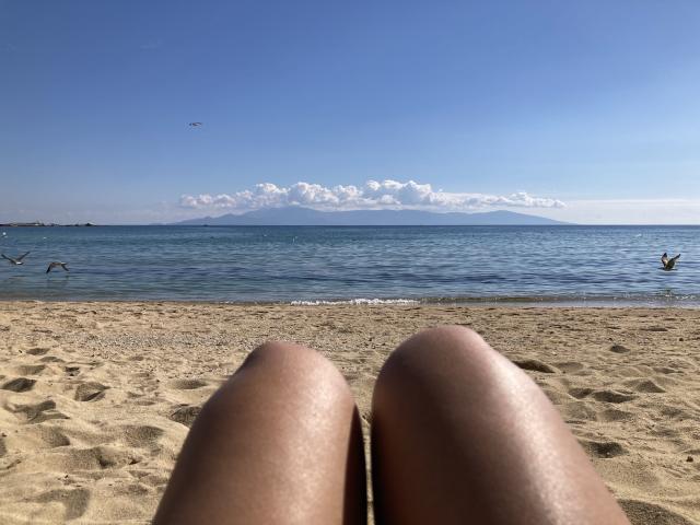 DeesseBleue's sun-kissed thighs in the sand, blue ocean, and distant mountains with clear sky and a few birds in the air, photographed from a relaxed, beachside perspective.