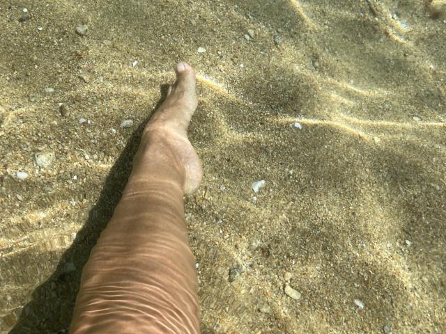 DeesseBleue's leg with a slightly tanned, brown-skinned foot standing in clear, shallow water, showing gentle ripples and small white pebbles on the sandy bottom.