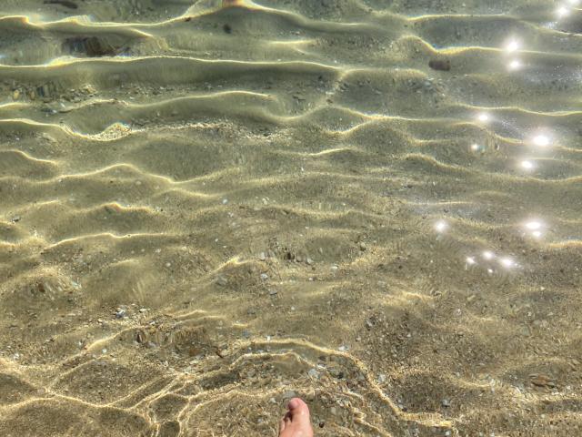 DeesseBleue's foot is in clear, rippling water with sunlight reflections, showing a sandy bottom with small rocks. The water's surface has wavy patterns and sparkling light.