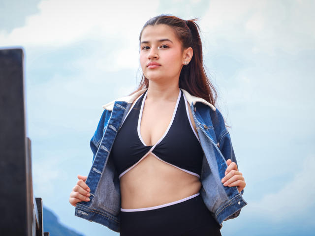 IrisBlair with brown ponytail, wearing black sports bra with cutout and denim jacket, showing her tits and flat stomach, standing with hands on jacket, looking confident.