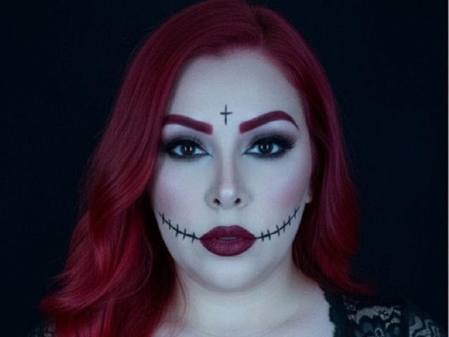 CatheKinkySwitch has red hair, dark eyeliner, and a black stitched mouth with lipstick. She has a black cross above her eyebrow and a dark, gothic makeup style.