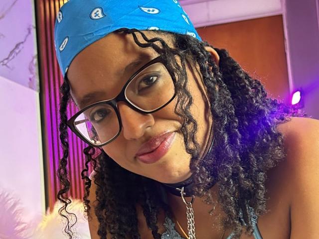 Artemisa, met krullend zwart haar, een blauwe bandana, zwarte bril, roze lippenstift en een zwarte choker met een ketting, glimlacht licht. Borsten niet zichtbaar, focus op het gezicht..