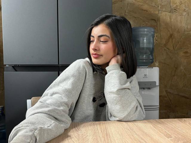 EmmaBeaumont with closed eyes, wearing a gray hoodie, has black shoulder-length hair, and rests her head on her hand, sitting at a wooden table, near a water cooler.