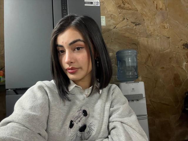 Emma Beaumont, avec son visage adorable, sa coupe de cheveux courte noire et son pull gris avec un motif de chat, a les lèvres légèrement pincées et regarde l'appareil photo. Un distributeur d'eau se trouve derrière elle..
