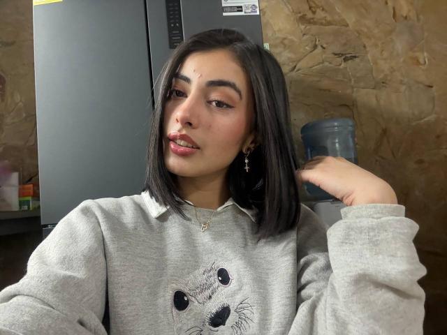 EmmaBeaumont with shoulder-length black hair, wearing a grey hoodie with a cat face, small nose ring, and earring, slightly smirking, hand near head, in a casual setting.