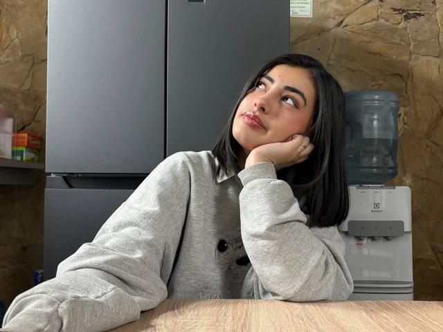EmmaBeaumont with shoulder-length black hair, wearing a grey sweater, head resting on hand, looking to the side with a slight smile, next to a water cooler and fridge.