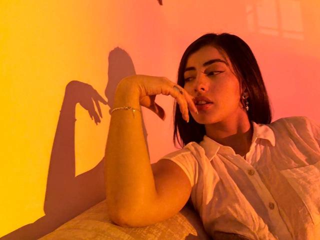 EmmaBeaumont with dark hair, wearing a white button-up shirt, resting her arm on a pillow, casting a shadow, glowing in warm yellow-orange light, looking relaxed and slightly sultry.