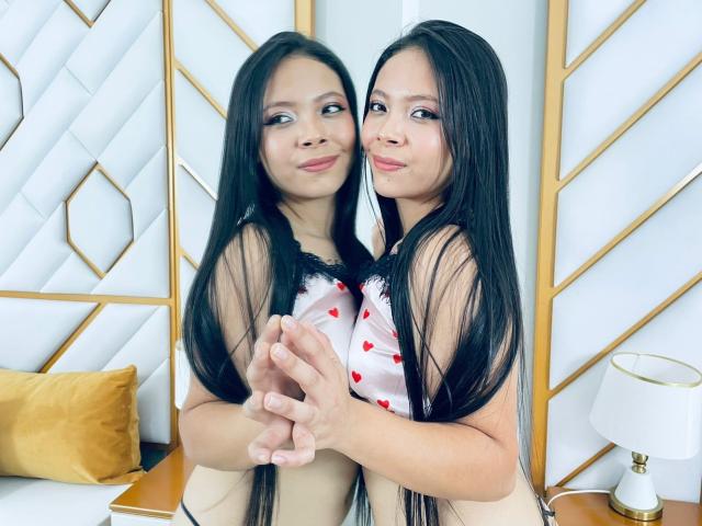 AnnieHill has long black hair, wearing a white top with red heart patterns, holding hands with her mirrored reflection. Her tits are small, she's smiling, and has light skin.