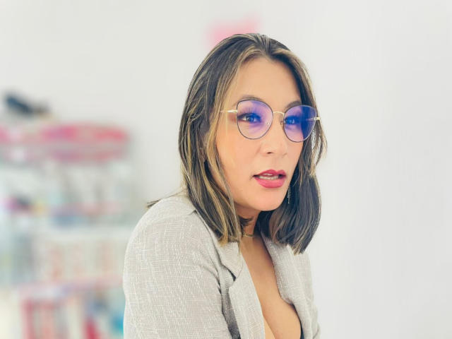 EmaFoxy, con un seno di media grandezza, indossa un blazer grigio, occhiali rotondi con montatura viola e rossetto rosso, ha i capelli castani che le arrivano alle spalle e un leggero sorriso, in una stanza luminosa..