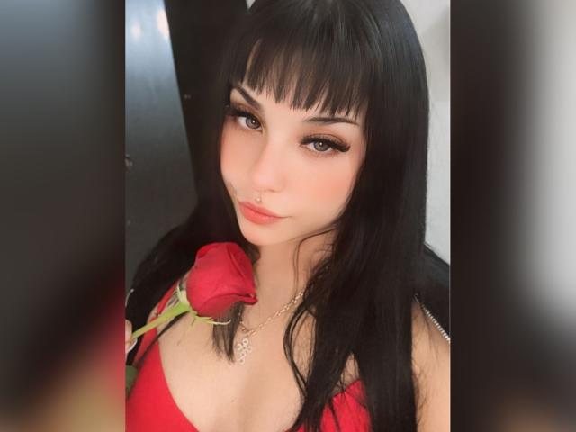 GraceClarke with black hair, bangs, and red lipstick, holding a rose against her chest, wearing a red top, and a nose ring, looking at the camera.