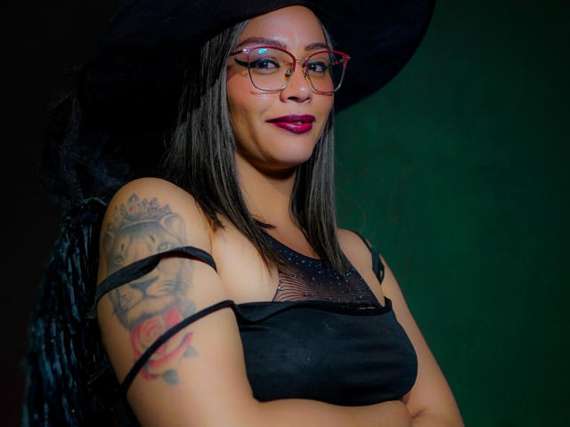 VeronicaSaienz with dark hair, red glasses, black hat, and black dress showing tits through mesh, arms crossed, tattoo on right arm, green background, and red lipstick.