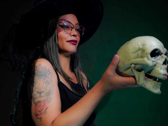 VeronicaSaienz with glasses, black dress, and witch hat, holding a skull, has a lion and rose tattoo on her arm, purple lipstick, and long dark hair.