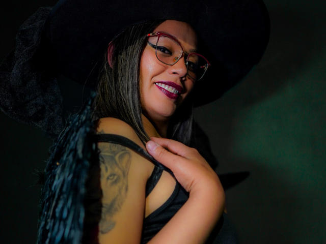 VeronicaSaienz with glasses, black witch hat, and black dress, smiling, showing tattooed arm, holding dress strap, dark green and black lighting, visible purple lipstick, and black wings.