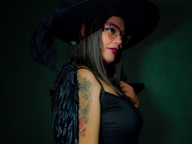 VeronicaSaienz in a witch outfit with a black hat, glasses, and fringed black vest, showing tattooed arm, with pink lipstick, looking sideways. Boobs, arm tattoo, dark green background.