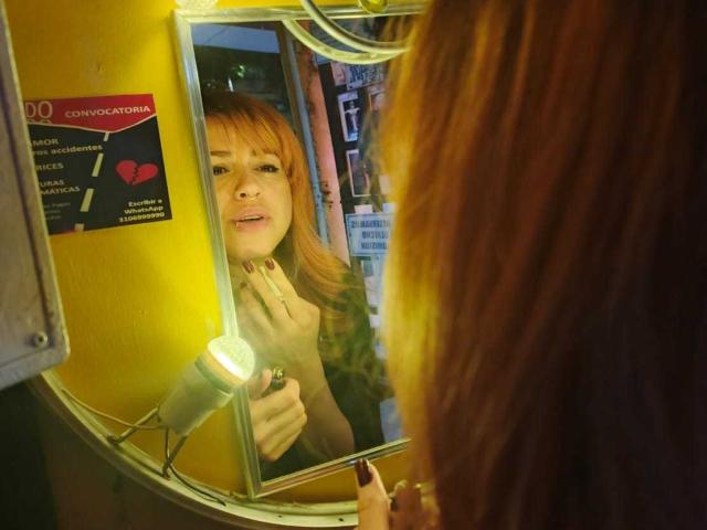 PaulettWatsoon with red hair, looking into a round mirror, holding her finger to her lips, reflection shows her face, yellow wall, mirror light on, nails painted dark.